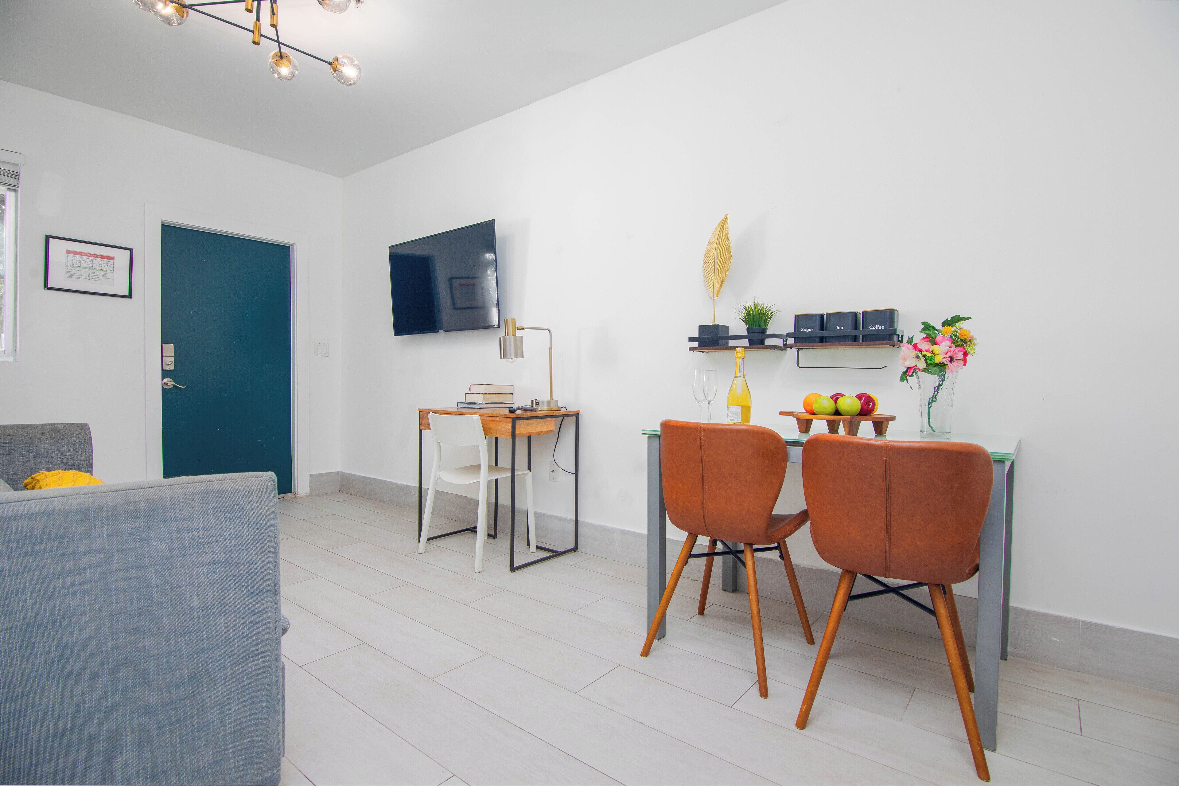 A modern living room with a sofa, a dining table with chairs, a TV mounted on the wall, and a small shelf with decorative items.