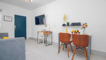 A modern living room with a sofa, a dining table with chairs, a TV mounted on the wall, and a small shelf with decorative items.