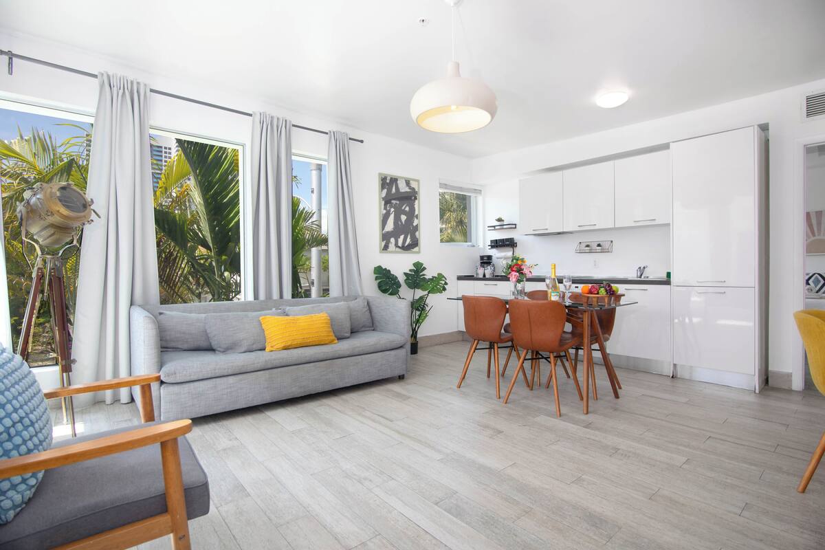 Ein modernes Wohnzimmer mit grauem Sofa, einem Holzespeisetisch und einer Küchenzeile mit weißen Schränken.