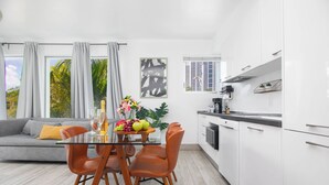 A modern kitchen with white cabinets, a dining area with a glass table and orange chairs, and a grey sofa with a yellow pillow. - Boutique Apartments SoBe (Miami Beach)