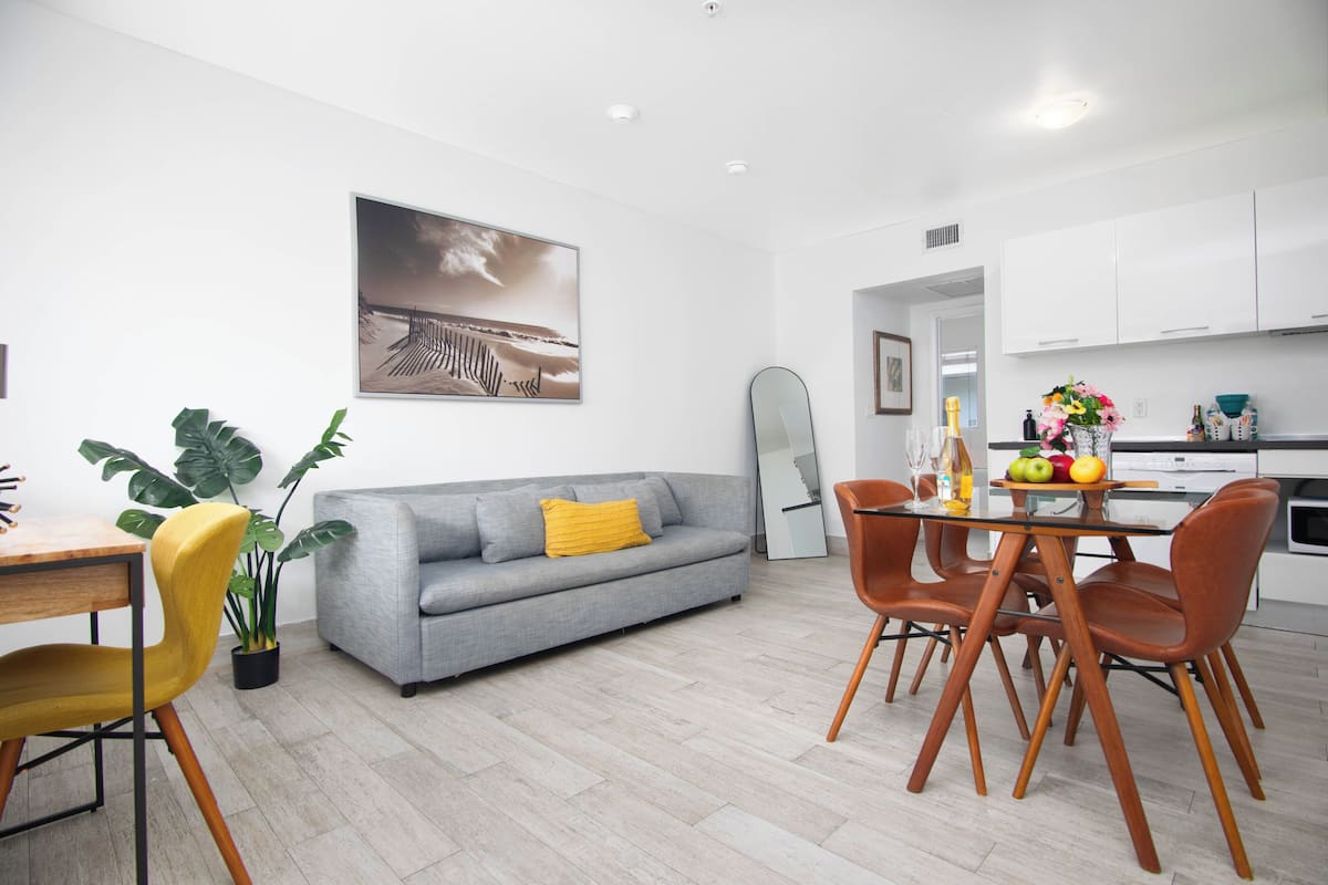 Ein moderner Wohnraum mit grauem Sofa, gelbem Sessel, Esstisch mit orangefarbenen Stühlen und einer Küchenzeile.