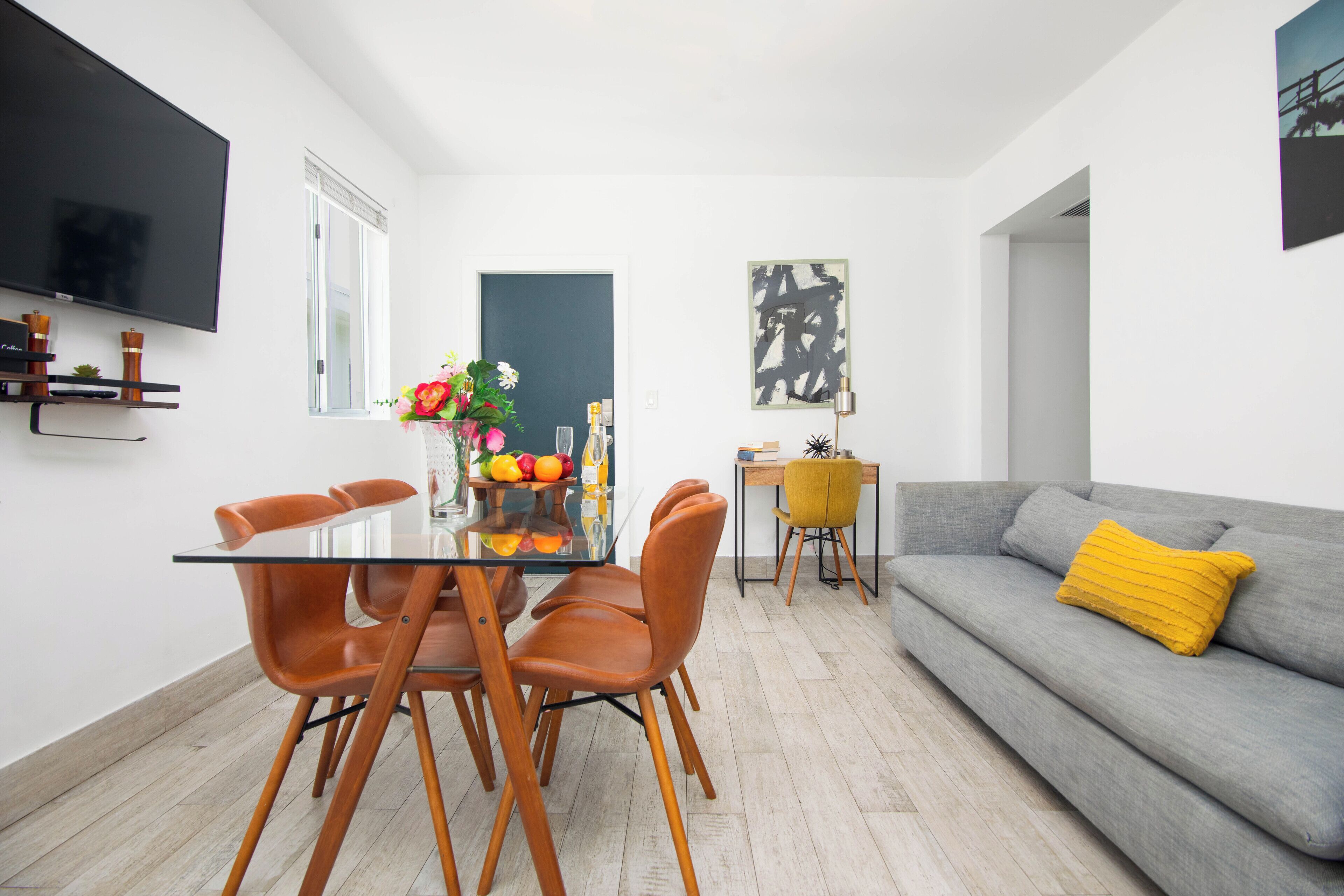 Un salon moderne avec un canapé gris, des fauteuils orange, une table à manger en verre et un téléviseur fixé au mur.