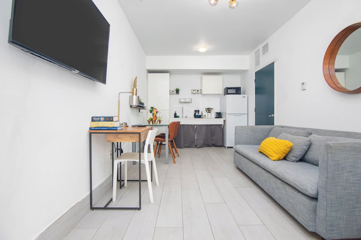 Ein modernes Wohnzimmer mit grauem Sofa, einem Esstisch mit Stühlen, einer Küchenzeile und einem Wand-Fernseher.