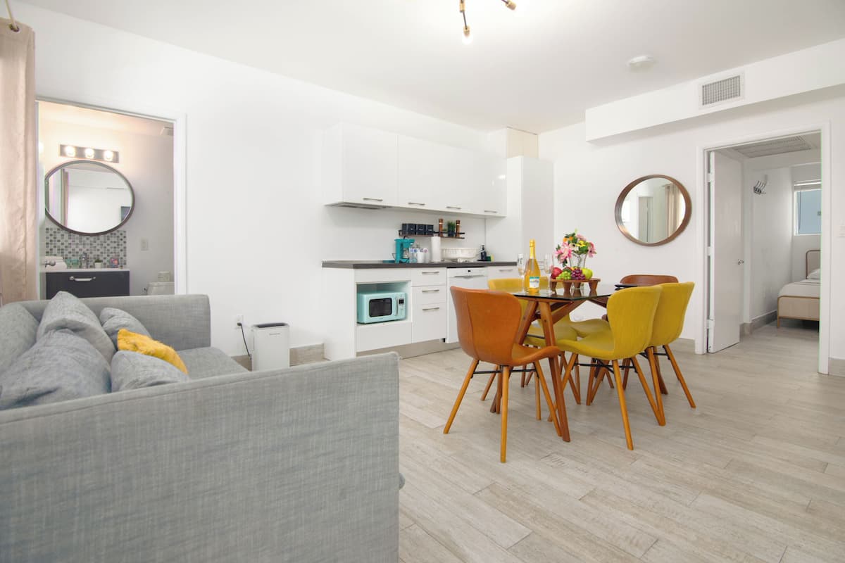 Ein modernes Wohnzimmer mit grauem Sofa, einem Esstisch mit gelben Stühlen und einer Küche mit weißen Schränken und einer Mikrowelle.