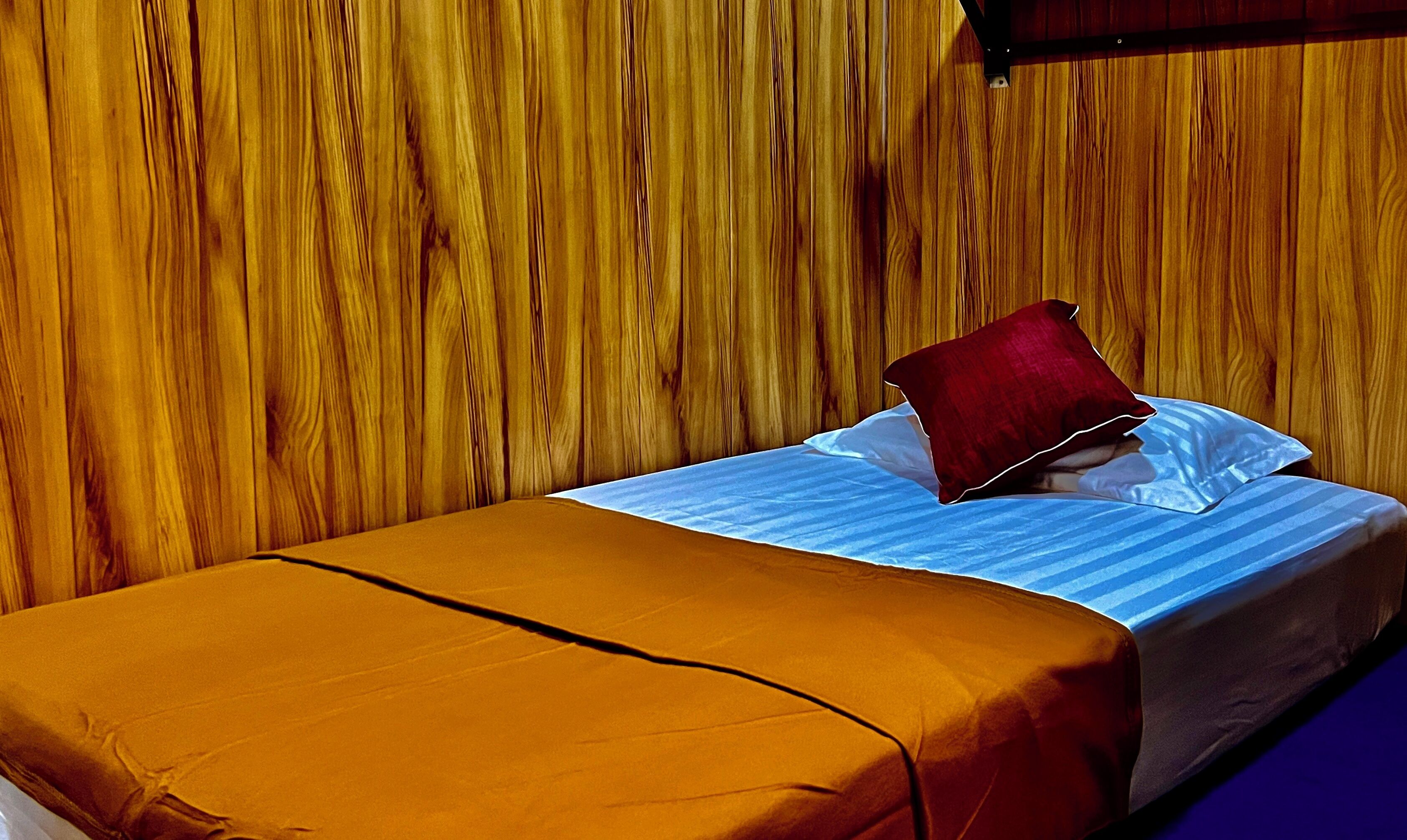 A single bed with a striped blue and white sheet, a red pillow, and a brown blanket. The room has wooden paneling on the walls.