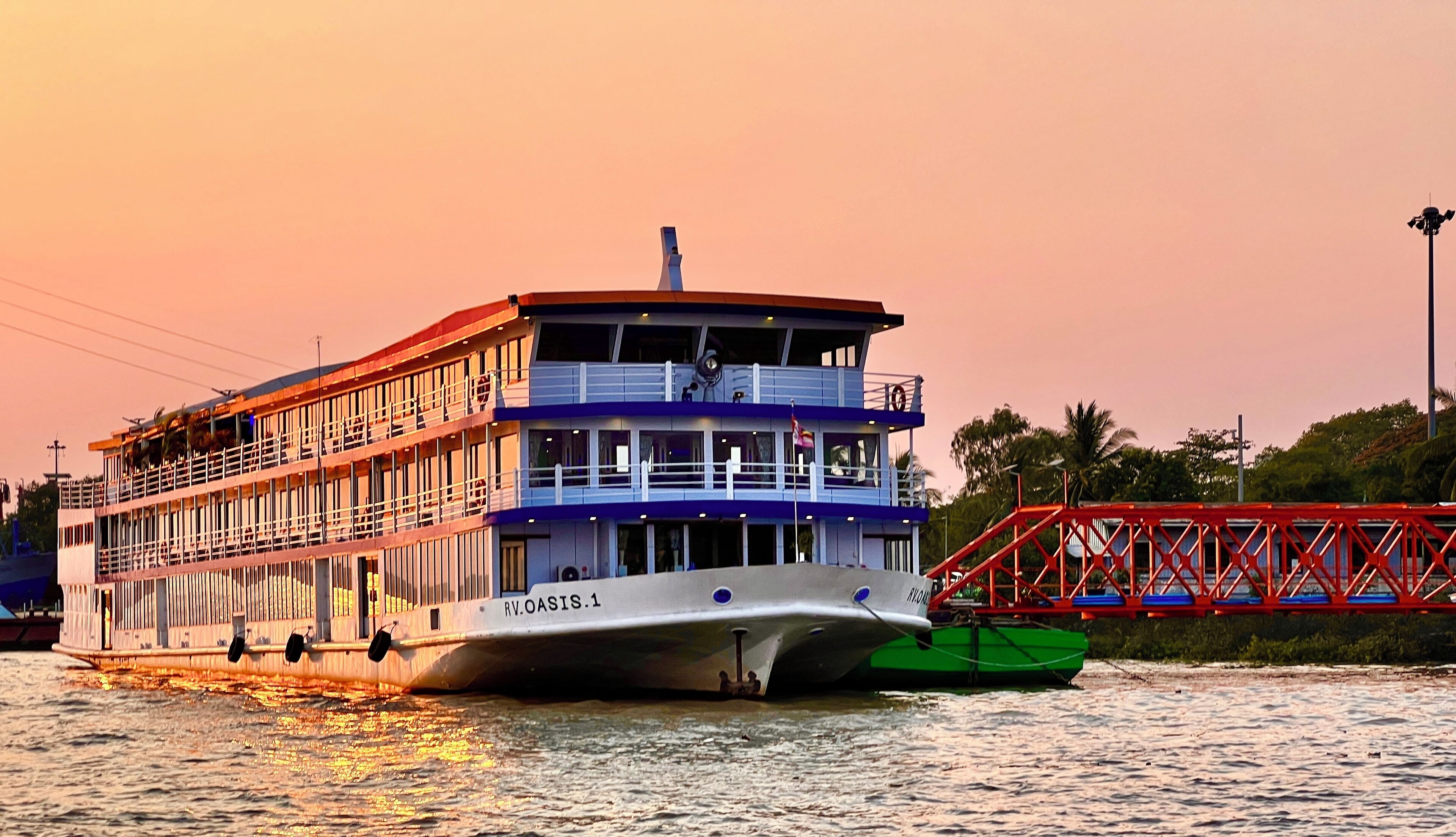 A multi-deck riverboat with a white and blue color scheme, named RV OASIS.1, docked at a pier with a red bridge in the background.