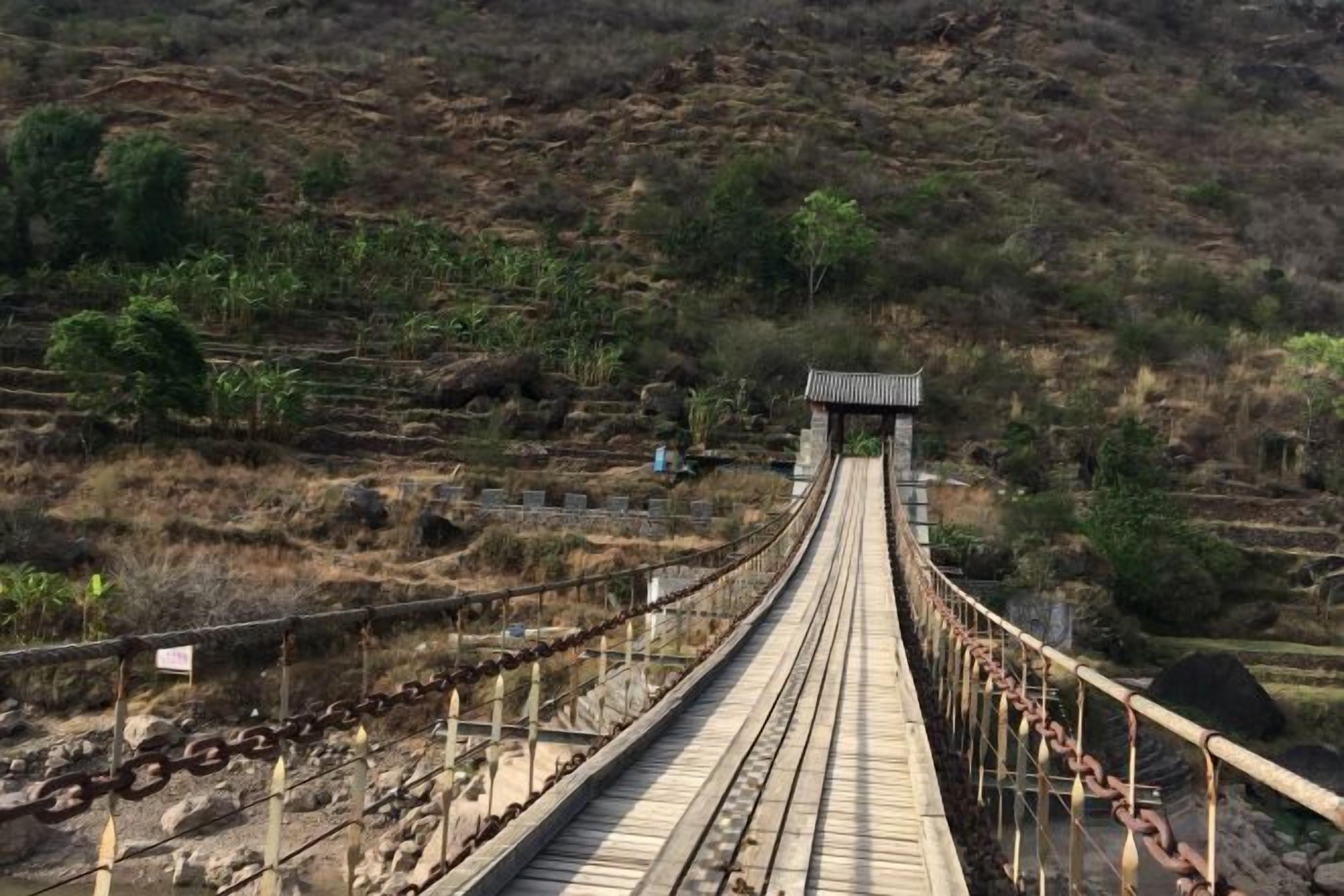 A long suspension bridge with a wooden walkway, surrounded by dry vegetation and a hilly landscape.