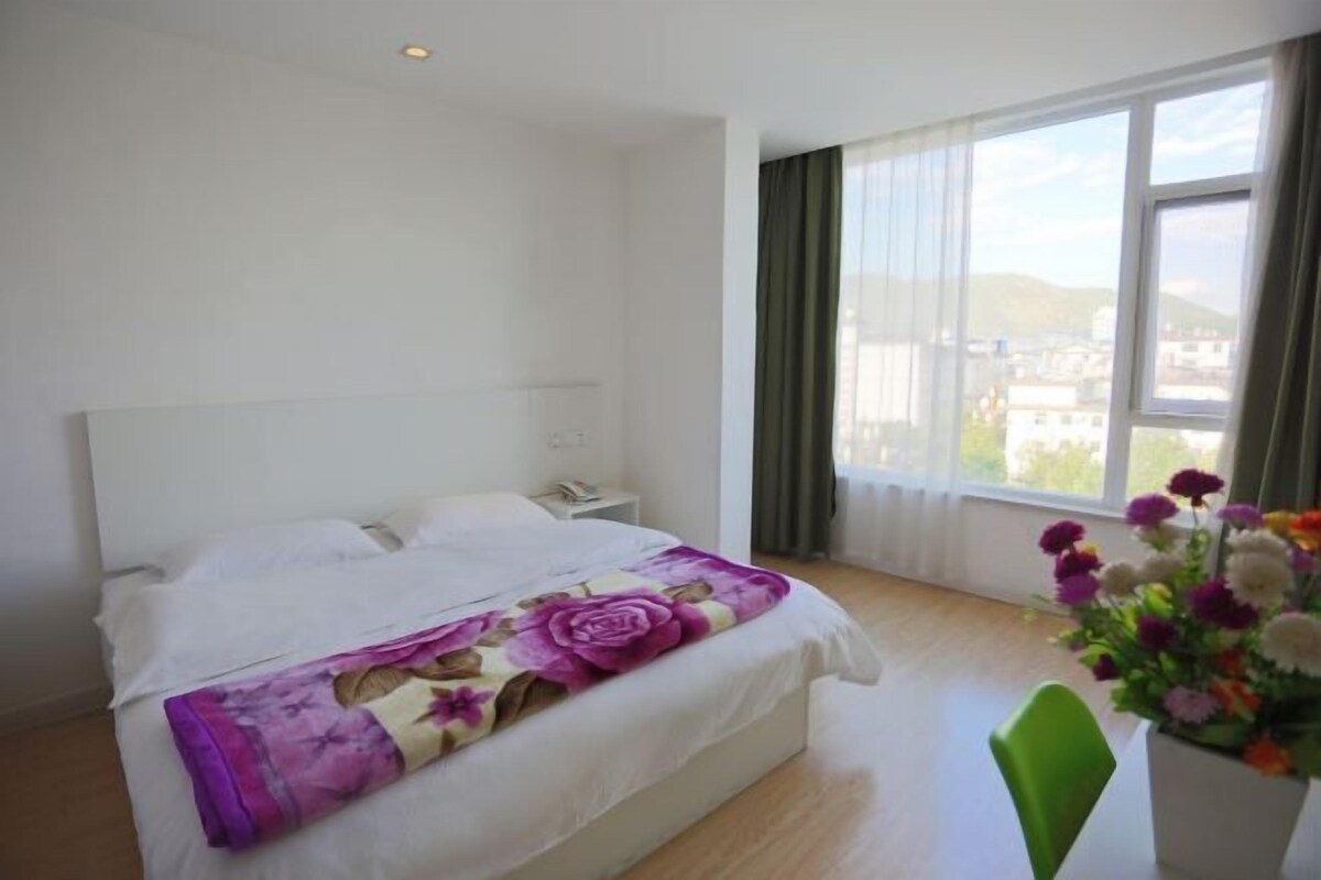 A hotel room with a bed, a nightstand, a chair, a window with curtains, and a flower vase.