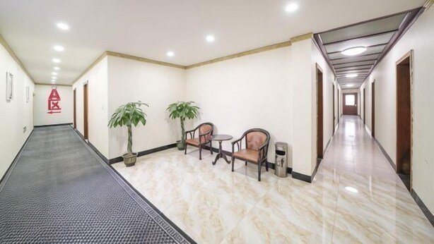A hotel corridor with a tiled floor, a seating area with chairs and a table, potted plants, and a trash bin.