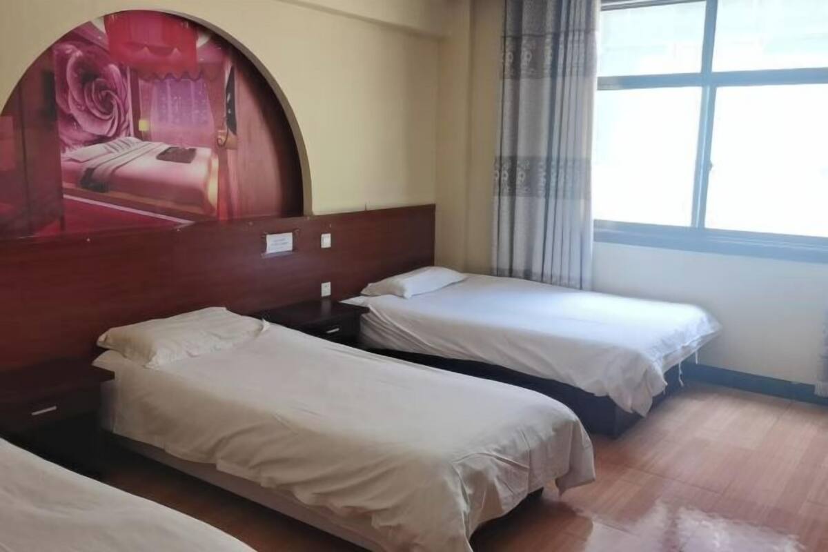 Een hotelkamer met drie enkele bedden, een houten hoofdeinde, een groot raam met gordijnen en een wanddecoratie.