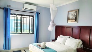 Une chambre d’hôtel avec un lit, une table de chevet, une fenêtre avec des rideaux, un appareil de climatisation et un tableau encadré au mur.