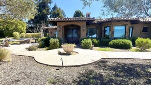 A Mediterranean-style house with a tiled roof, a central entrance with a decorative door, and a landscaped garden with a large planter and trimmed bushes. - Luxury Home on Secluded Ranch, near beaches and hiking (San Diego)