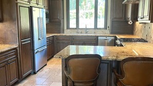 A spacious kitchen with dark wood cabinets, a stainless steel refrigerator, a large island with bar stools, and a window providing natural light. - Luxury Home on Secluded Ranch, near beaches and hiking (San Diego)