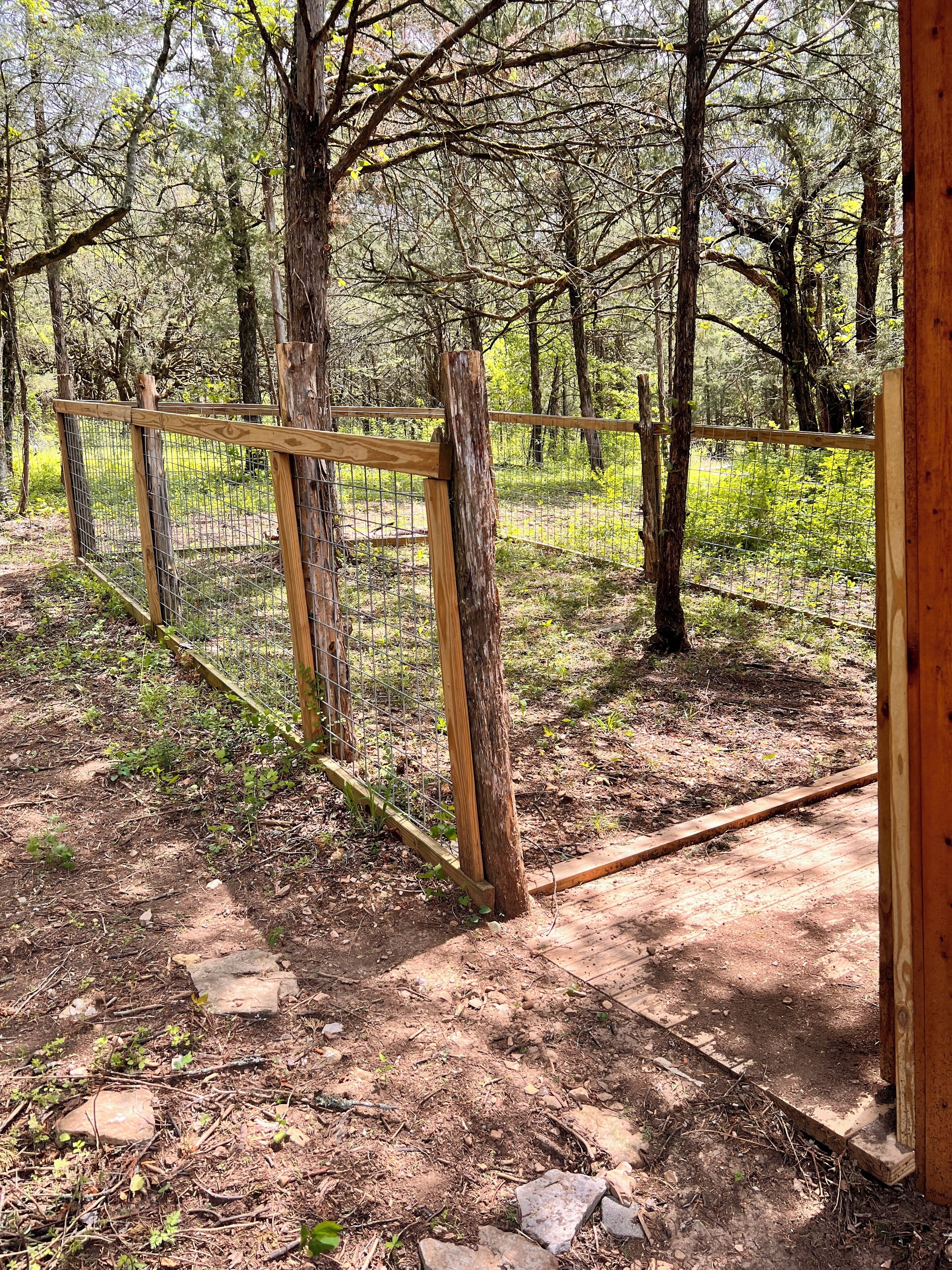 Espacio al aire libre cercado con una estructura de madera, rodeado de árboles y suelo de tierra.