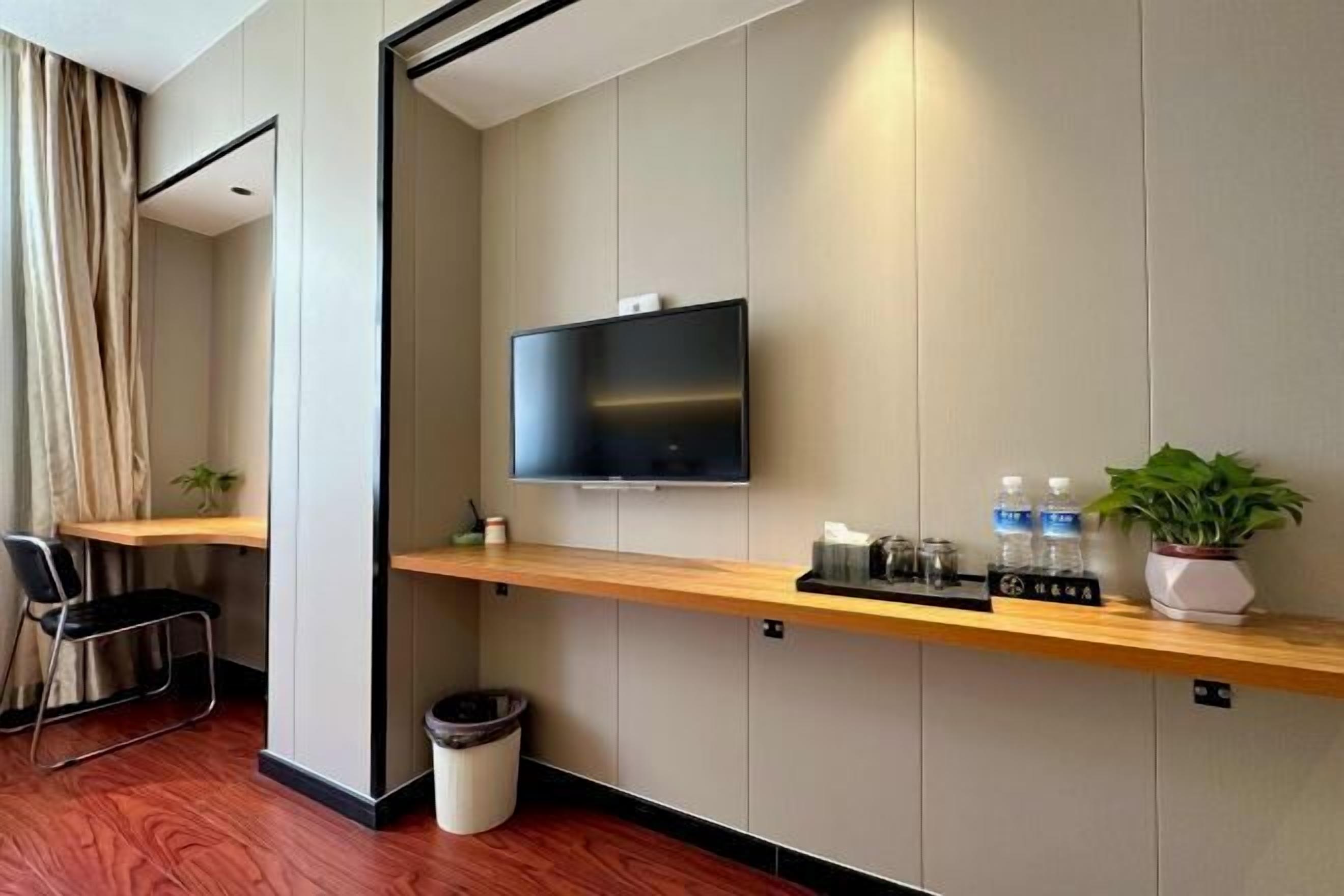 Una habitación de hotel moderna con un televisor de pantalla plana montado en la pared, un escritorio de madera, una silla, un bote de basura pequeño y una planta en maceta.
