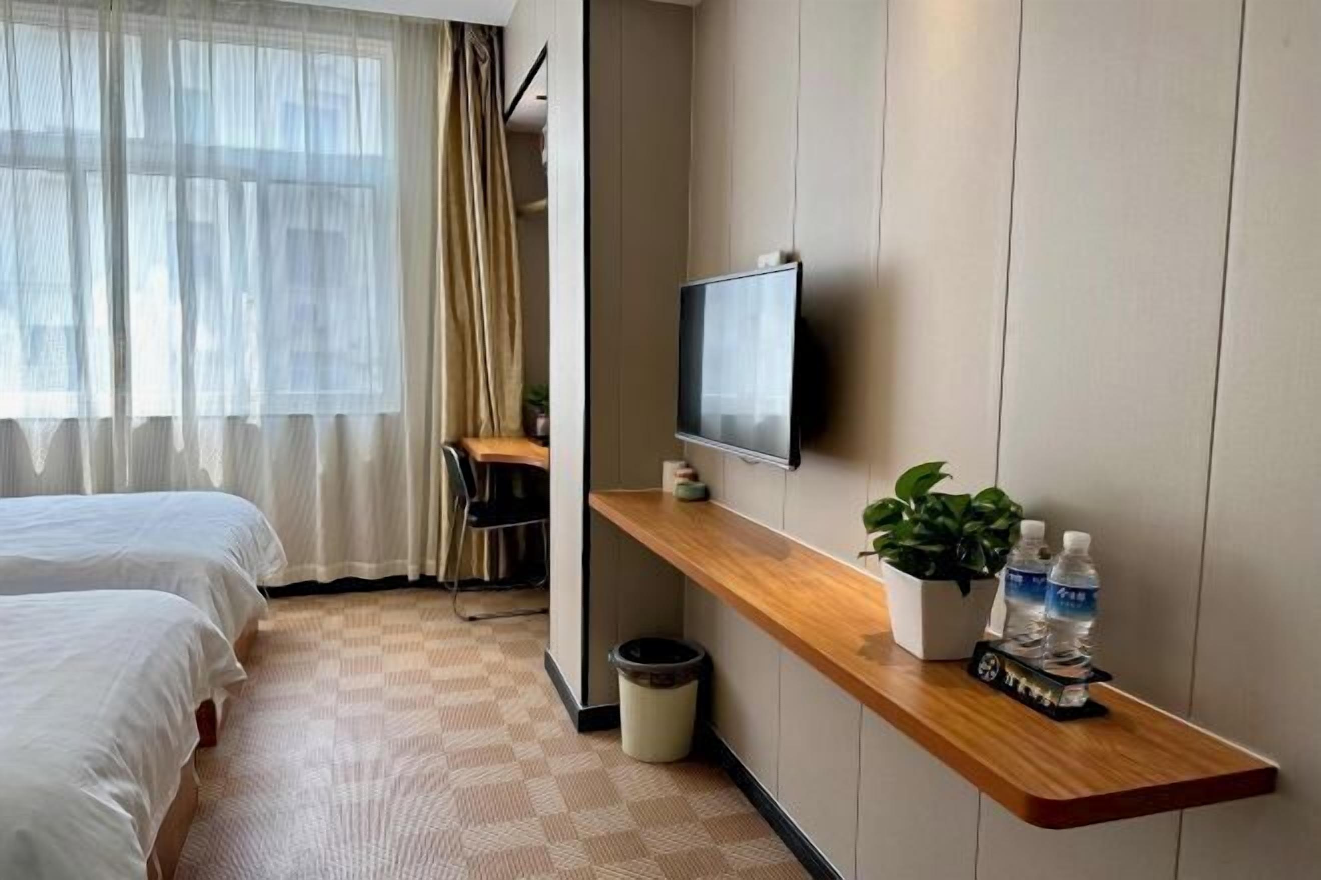 A hotel room with two beds, a desk, a TV, a shelf with a plant and water bottles, and a trash bin.