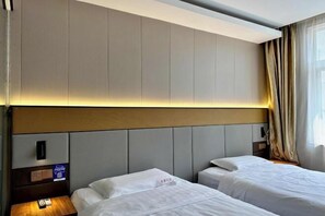 Habitació d'hotel amb dos llits, un capçal amb il·luminació integrada, una taula de nit amb làmpada i una finestra amb cortines.