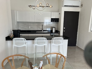 A modern kitchen with white cabinetry, a black countertop, and a built-in coffee station. There are three white chairs around a glass table.