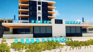 A modern hotel with a multi-story structure, a pool area with blue lounge chairs, and a clear blue sky.