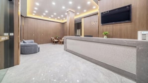 Zona de recepción de un hotel con piso de mármol, un mostrador de recepción, un televisor de pantalla plana y un área de descanso con una mesita y sillas.