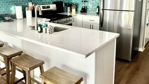 A modern kitchen with white cabinets, a stainless steel refrigerator, a large island with stools, and a teal backsplash. - Family-friendly, Updated Condo- Stocked Beach Closet-Steps to Crescent Beach-NMB (North Myrtle Beach)