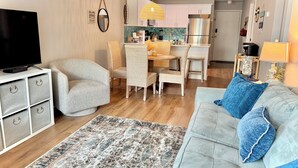 A modern living room with a grey sofa, blue cushions, a flat-screen TV on a white cabinet, wicker chairs, a patterned rug, and a kitchen area in the background. - Family-friendly, Updated Condo- Stocked Beach Closet-Steps to Crescent Beach-NMB (North Myrtle Beach)