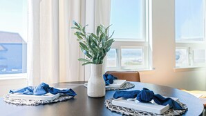 A dining table with a white textured vase containing green leaves, blue napkins, and a white rectangular object on a woven placemat. - 2 Cayucan Bungalows Steps to Beach + Shared Lounge Sleeps 12 (Cayucos)