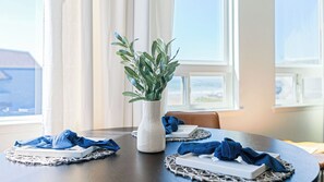 A dining table with a white textured vase containing green leaves, blue napkins, and a white rectangular object on a woven placemat. - 2 Cayucan Bungalows Steps to Beach + Shared Lounge Sleeps 12 (Cayucos)