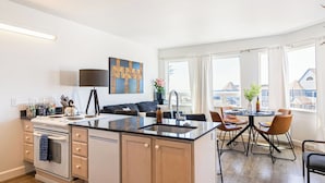 A modern kitchen with a central island, a dining area with a round table, and large windows offering a view of buildings. - 2 Cayucan Bungalows Steps to Beach + Shared Lounge Sleeps 12 (Cayucos)