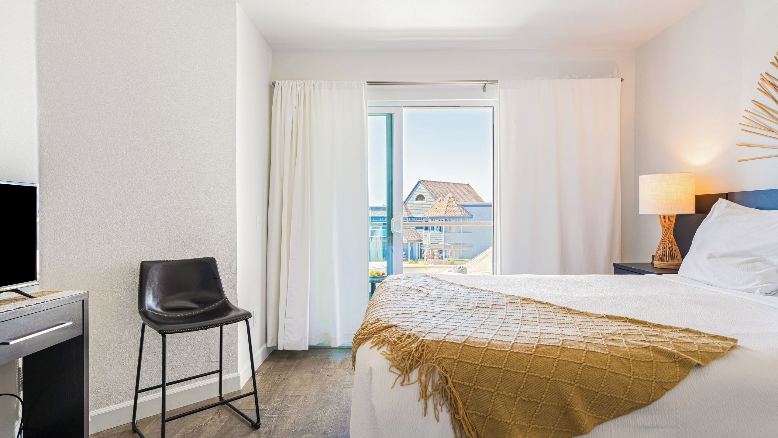 A hotel room with a bed, a desk with a chair, a window with curtains, and a view of buildings outside.
