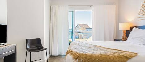 A hotel room with a bed, a desk with a chair, a window with curtains, and a view of buildings outside.