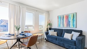 A modern living room with a blue sofa, a dining table, and a large window with curtains. - 2 Cayucan Bungalows Steps to Beach + Shared Lounge Sleeps 12 (Cayucos)