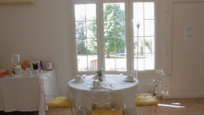 Un coin repas avec une table ronde, une nappe blanche et des chaises rembourrées de couleur jaune. Une fenêtre offre une vue sur la verdure à l’extérieur.