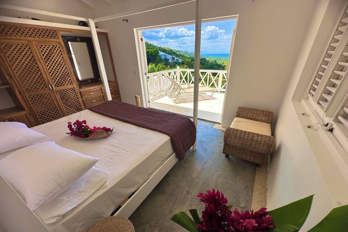 A bedroom with a bed, a small seating area, a balcony with a view of the sea, and a wooden wardrobe.