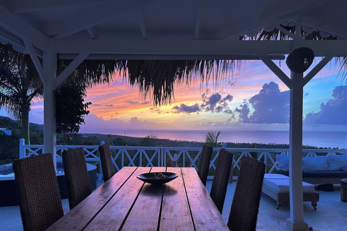 A wooden table with chairs under a white pergola, overlooking a scenic sunset view.