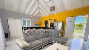 A modern living room with a grey sectional sofa, a white coffee table, and a kitchen area with appliances and a yellow wall. - Perrin Évasion 1CH à 9 min de l'aéroport de la Guadeloupe (Les Abymes)