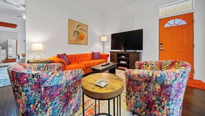 Una sala de estar con un sofá naranja, sillones coloridos, una mesa de centro, un televisor y un cuadro en la pared.