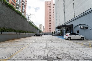 A parking area with two cars, a building with a blue door, and a tall pink building in the background. - Perdizes Residence Flat Condomínio  (São Paulo)