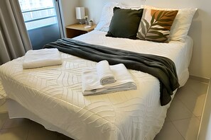 Una cama bien hecha con sábanas blancas y un plaid oscuro, una mesita de noche con una lámpara y una planta pequeña, y una ventana con cortinas.