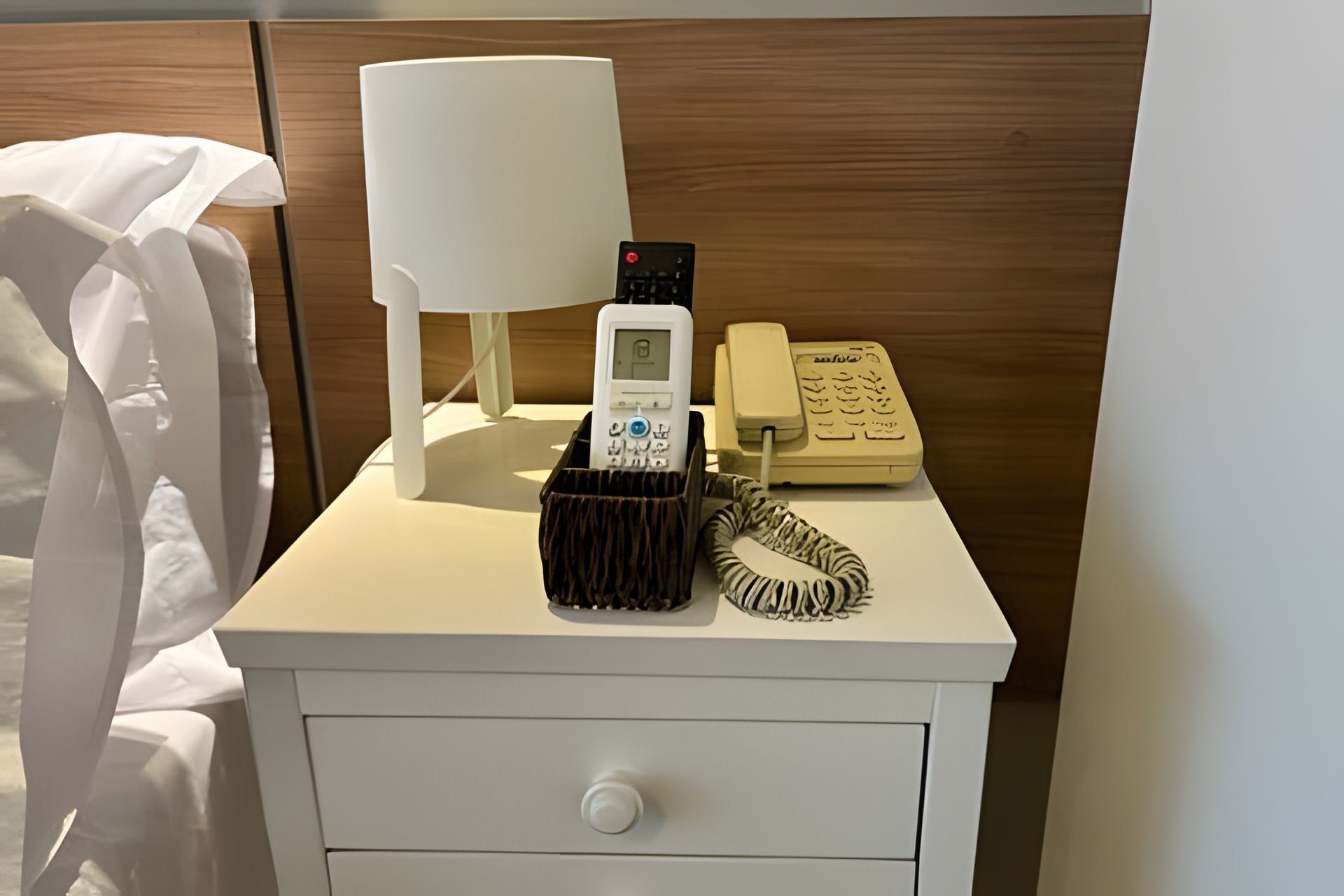 A white bedside table with a lamp, a telephone, a telephone charger, and a drawer.