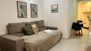 A neutral-toned living area with a sofa, a small table, and a chair. There are framed artworks on the wall, a light fixture, and a glimpse of a kitchen area.