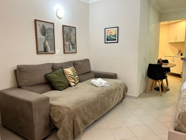 A neutral-toned living area with a sofa, a small table, and a chair. There are framed artworks on the wall, a light fixture, and a glimpse of a kitchen area. - Perdizes Residence Flat CondomÃnio (São Paulo)