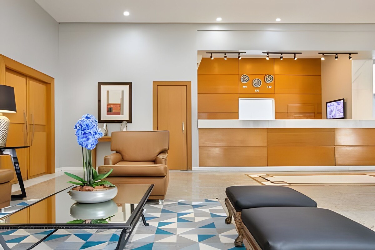 Uma área de recepção moderna com balcão, sofá, poltrona, mesa de centro com uma planta e piso estampado.