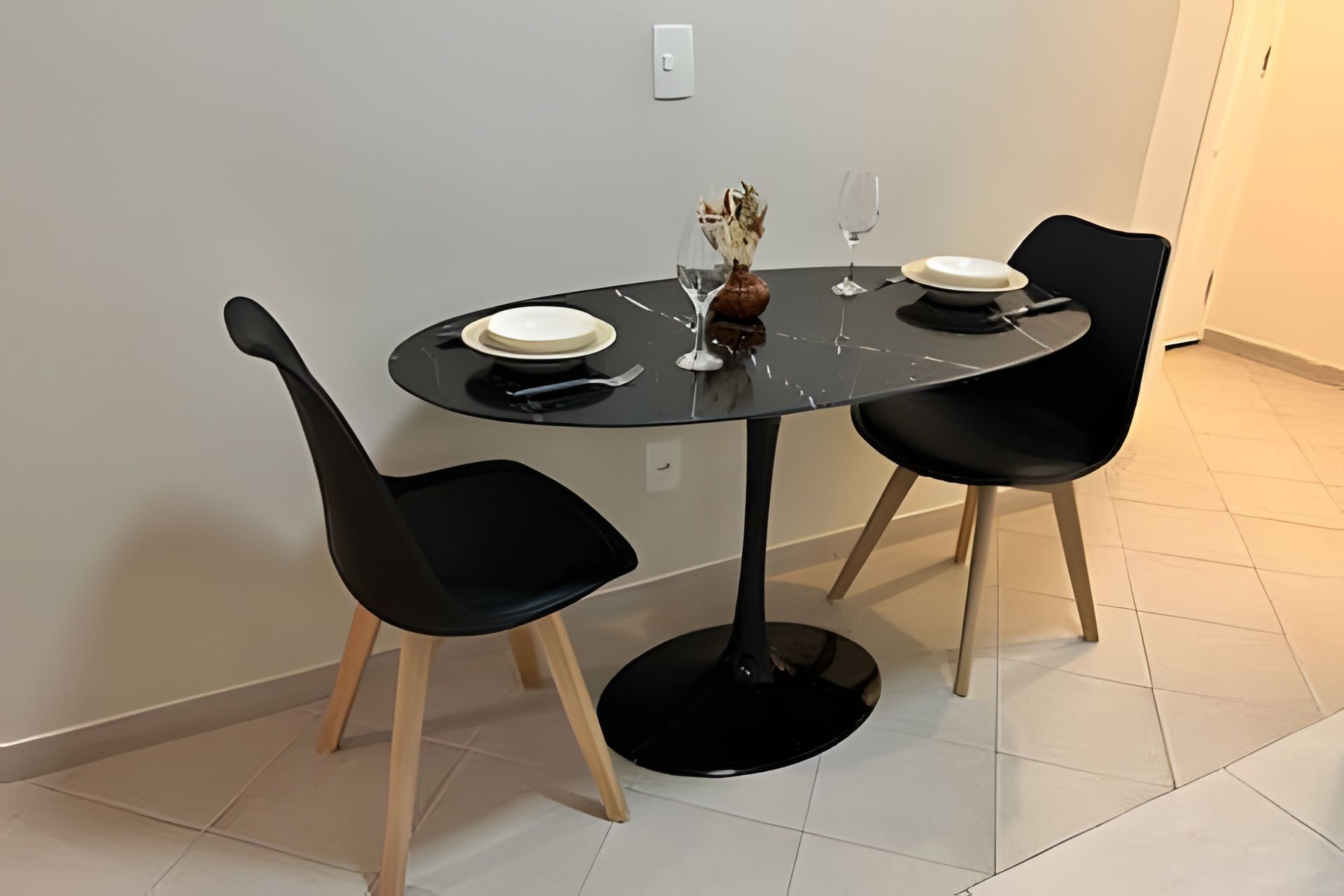 Uma mesa de jantar redonda com cadeiras pretas, um vaso com flores secas e dois copos de vinho.