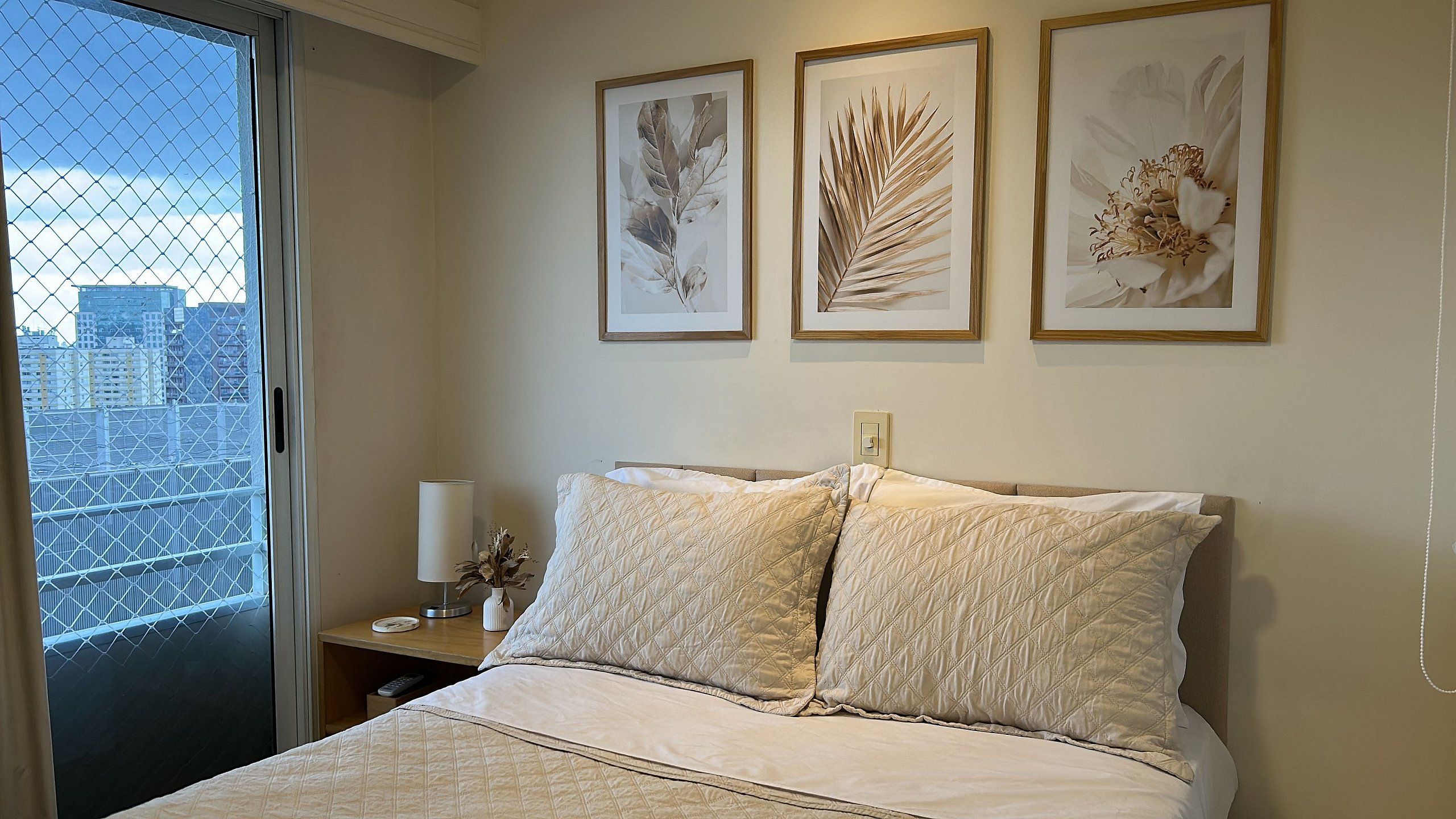 Habitación de hotel con cama, mesita de noche, lámpara y tres láminas botánicas enmarcadas en la pared.