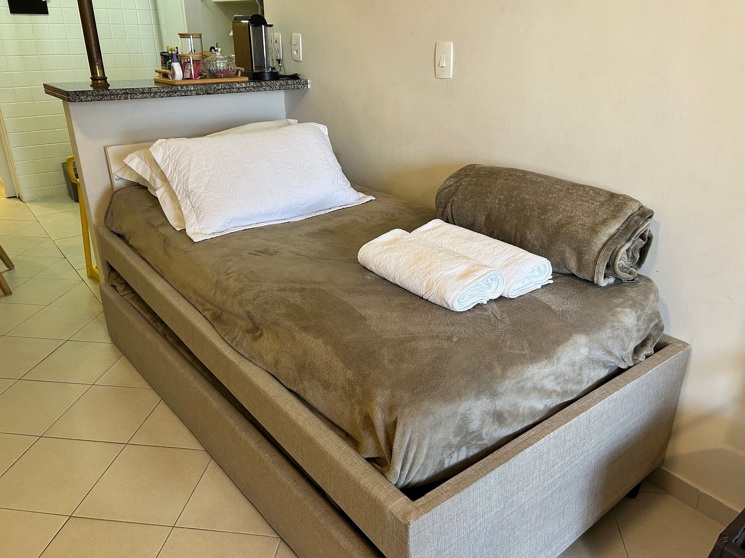 A single bed with a brown cover, a white pillow, and two rolled towels.