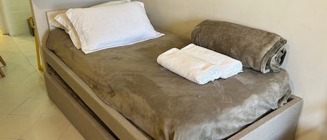 A single bed with a brown cover, a white pillow, and two rolled towels.