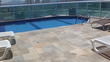 A rooftop pool with a glass balustrade, stone tiles, and white lounge chairs.