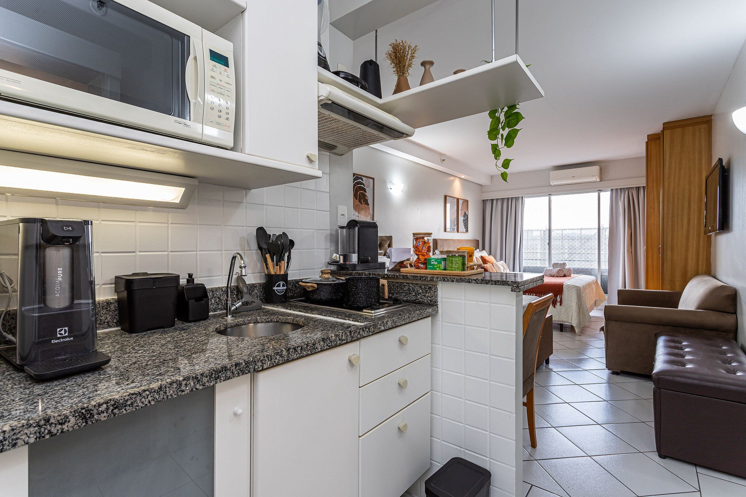 A modern kitchen with a microwave, coffee maker, and a sink. There is a dining area with a sofa and a TV.