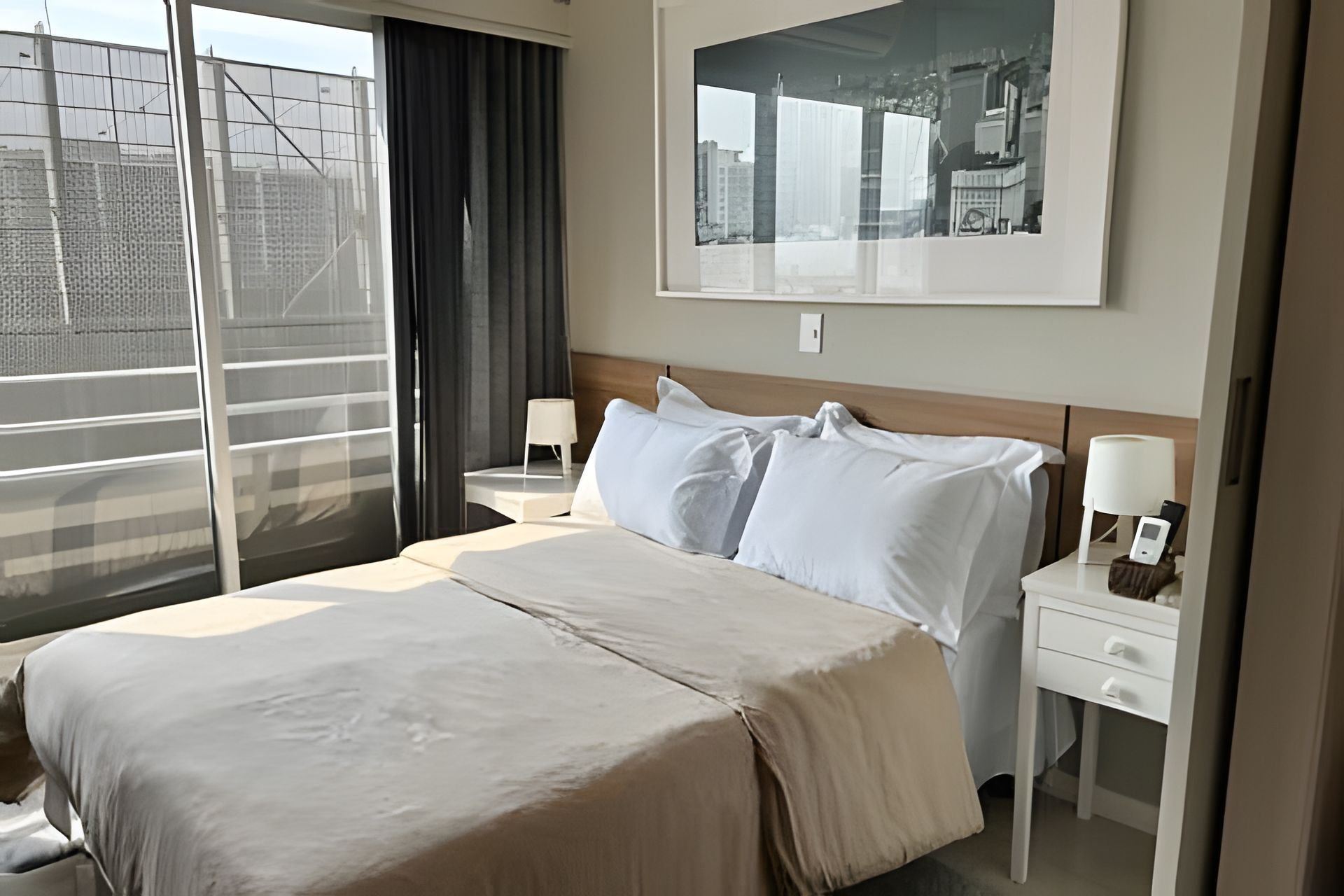 A hotel room with a large bed, two bedside tables with lamps, a window with a view of buildings, and a framed picture on the wall.